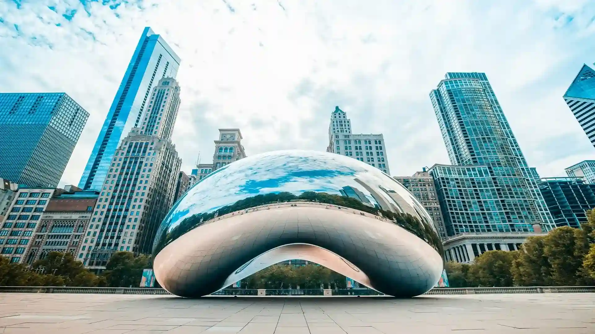 A large reflective sculpture known as "The Bean" in Chicago's Millennium Park, surrounded by tall buildings under a partly cloudy sky